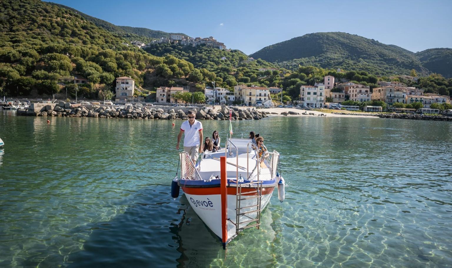 Barca turistica in navigazione nelle acque cristalline, con un pittoresco borgo costiero circondato da colline verdi sullo sfondo.