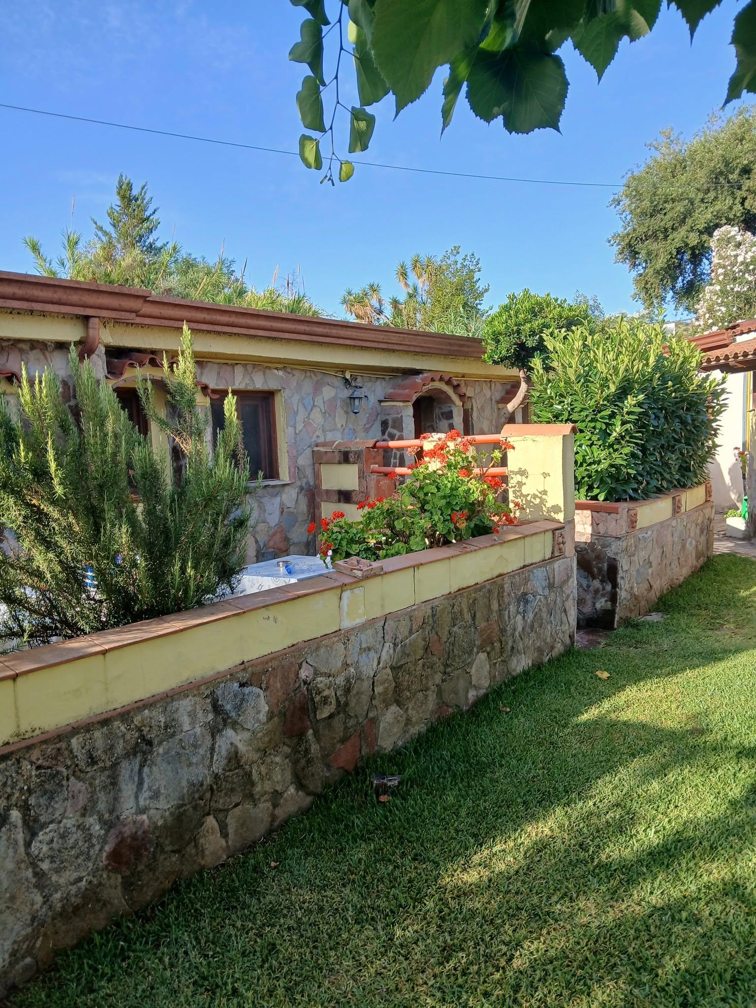 Casa in pietra con giardino curato, aiuole fiorite e muretto rustico in un ambiente verde e soleggiato.
