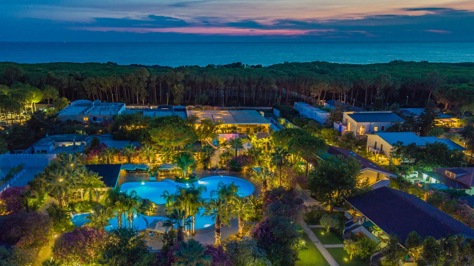 Vista aerea del resort immerso nel verde, con piscina illuminata e mare sullo sfondo al tramonto.