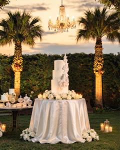 Torta nuziale bianca a tre piani con rose, candele e lampadario in un elegante allestimento da matrimonio all’aperto al tramonto.