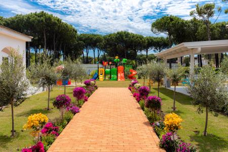 Ingresso dell'area giochi per bambini con vialetto in mattoni e aiuole fiorite colorate.
