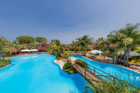 Piscina panoramica del resort con acqua turchese, palme, lettini e ombrelloni immersi nel verde.