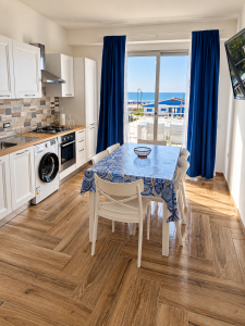 Cucina moderna e luminosa con tavolo da pranzo, tende blu e vista sul mare.