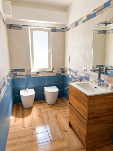 Bagno moderno con piastrelle beige e blu, mobile lavabo in legno, sanitari bianchi e pavimento effetto legno.